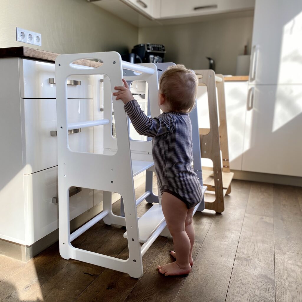 Viengadīgs bērns demonstrē virtuves palīgus. Attēlā redzami trīs krāsu varianti: balta, pelēka un koka krāsa.
