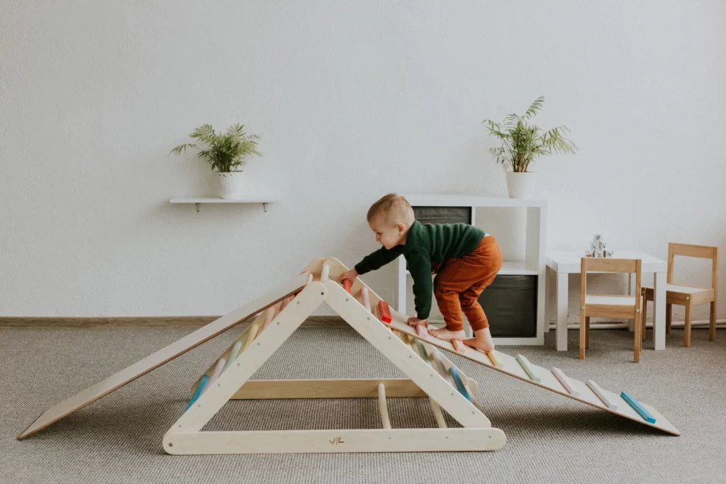 A climbing triangle made of natural wood that is diligently climbed by a child under the age of three.
